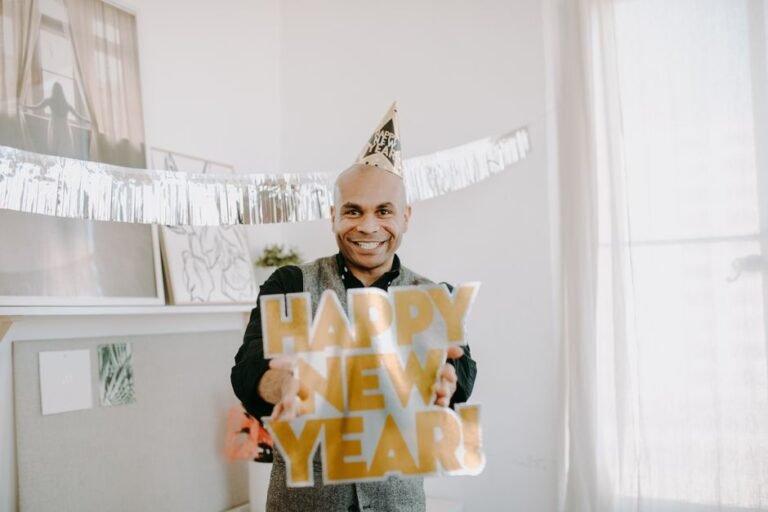 A smiling man celebrates New Year's indoors with a festive sign and party hat, filled with joy and warmth.
