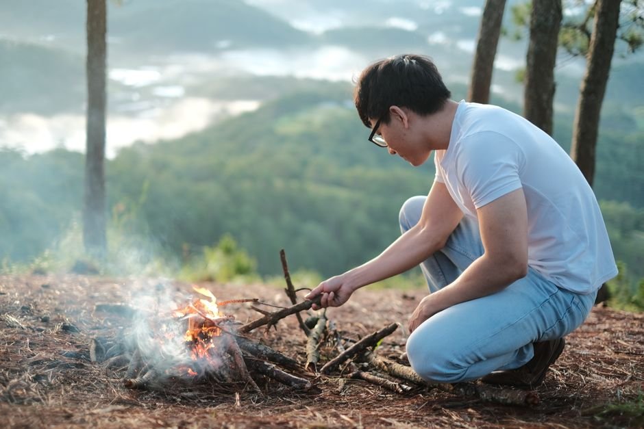 Young man in a white shirt tending a campfire on a hill with a scenic mountain view.
