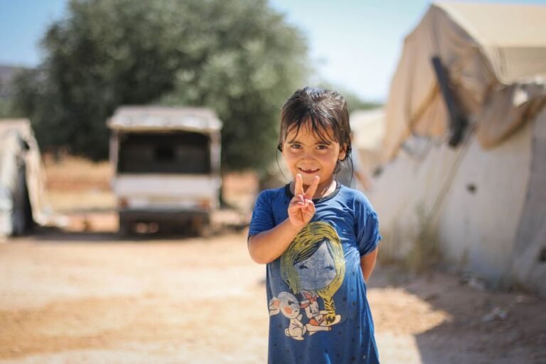 Young girl in Idlib, Syria, smiles and shows peace sign amidst tents.