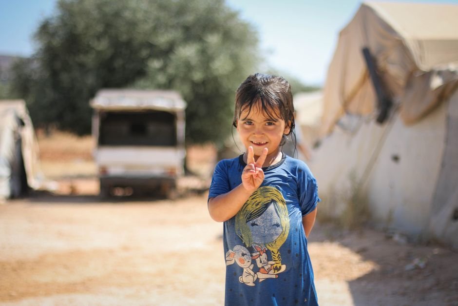 Young girl in Idlib, Syria, smiles and shows peace sign amidst tents.