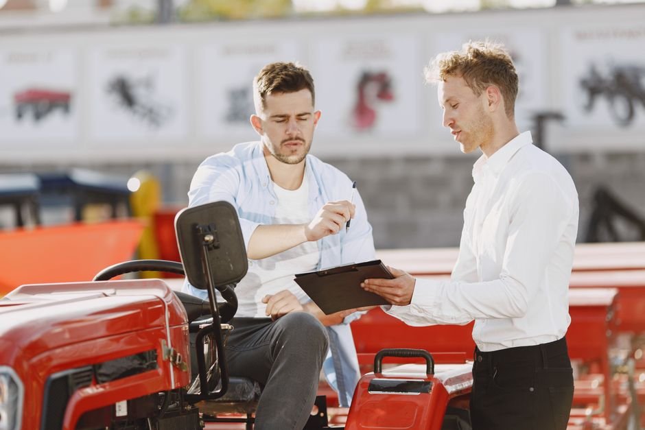 Two professionals discuss on a red tractor, examining paperwork under bright sunlight.