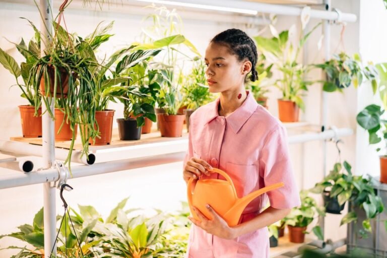 Woman in pink shirt holding watering can, surrounded by potted plants on shelves.