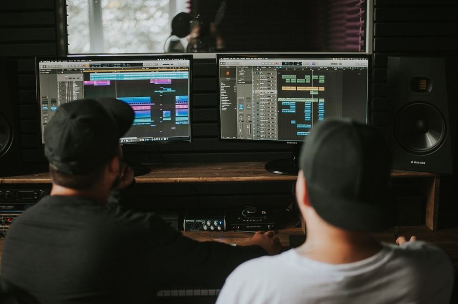 Two men working in a music studio with dual monitors displaying audio editing software.