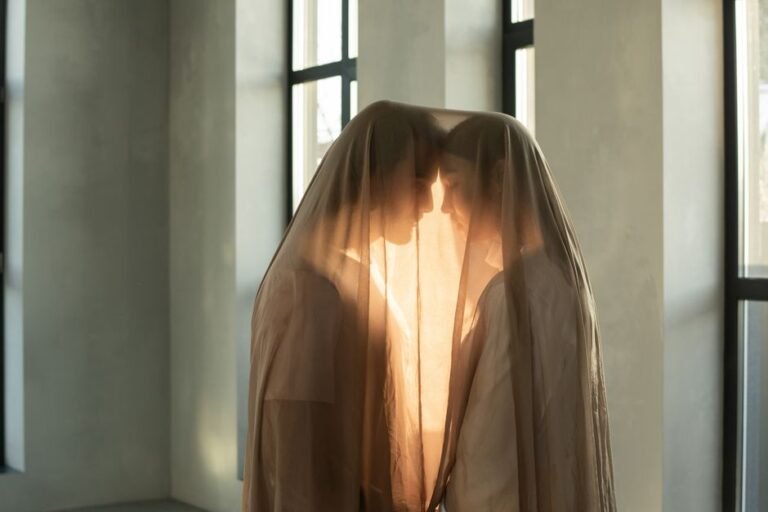 Two people in a romantic embrace under a sheer veil indoors, bathed in warm light.